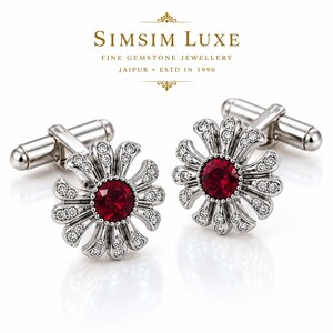 Puede incluir: Un par de gemelos plateados con diseño de flor, con una piedra roja redonda central rodeada de pequeñas piedras transparentes en cada pétalo. El texto SIMSIM LUXE FINE GEMSTONE JEWELLERY JAIPUR ESTD IN 1990 es visible en la parte superior.