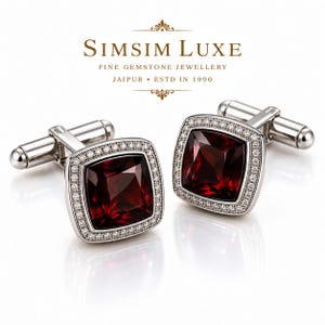 Puede incluir: Un par de gemelos de tono plateado con una piedra preciosa granate de color rojo intenso y corte cuadrado en el centro, rodeada por un borde de pequeñas piedras claras. El texto SIMSIM LUXE FINE GEMSTONE JEWELLERY JAIPUR ESTD IN 1990 es visible.