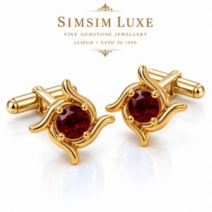 Op de afbeelding: Een paar gouden manchetknopen met een ronde, donkerrode edelsteen in het midden van een wervelend, vierpuntig metalen ontwerp. De tekst SIMSIM LUXE FINE GEMSTONE JEWELLERY JAIPUR ESTD IN 1990 staat boven de sieraden afgebeeld.