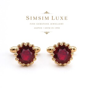 Könnte beinhalten: Ein Paar goldene Manschettenknöpfe mit runden, facettierten dunkelroten Edelsteinen, die von einem perlenartigen Goldrand umgeben sind. Darüber steht der Text SIMSIM LUXE FINE GEMSTONE JEWELLERY JAIPUR ESTD IN 1990 in brauner Serifenschrift.