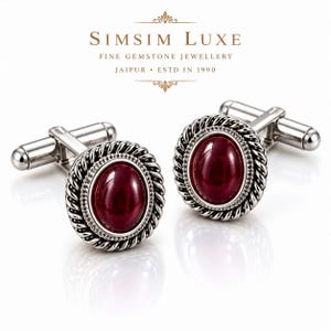 Op de afbeelding: Een paar zilverkleurige manchetknopen met een centrale ovaalvormige donkerrode edelsteen. De steen is omringd door een gedraaide touwachtige metalen rand en een kralenrand. De tekst SIMSIM LUXE FINE GEMSTONE JEWELLERY JAIPUR ESTD IN 1990 is zichtbaar.