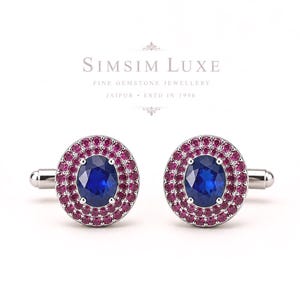Könnte beinhalten: Ein Paar silberfarbene Manschettenknöpfe mit einem zentralen, oval geschliffenen blauen Edelstein, umgeben von zwei konzentrischen Kreisen aus kleinen, runden rosa Edelsteinen. Der Text SIMSIM LUXE FINE GEMSTONE JEWELLERY JAIPUR ESTD IN 1990 steht darüber.