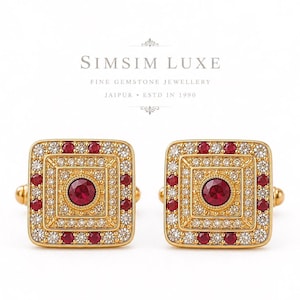 Op de afbeelding: Een paar vierkante gouden manchetknopen met een centrale ronde rode edelsteen omringd door concentrische randen van kleine witte steentjes en rode edelstenen. De tekst SIMSIM LUXE FINE GEMSTONE JEWELLERY JAIPUR ESTD IN 1990 staat boven de manchetknopen.