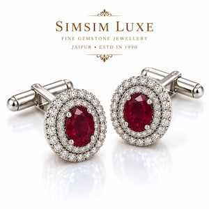 Könnte beinhalten: Ein Paar silberfarbene Manschettenknöpfe mit oval geschliffenen, tiefroten Edelsteinen, umgeben von zwei konzentrischen Ringen aus kleinen, klaren Steinen. Der Text SIMSIM LUXE FINE GEMSTONE JEWELLERY JAIPUR ESTD IN 1990 steht über den Manschettenknöpfen.