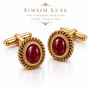 May include: A pair of gold-toned cufflinks featuring a central oval-shaped deep red gemstone surrounded by a twisted rope border and a beaded edge. The text SIMSIM LUXE FINE GEMSTONE JEWELLERY JAIPUR ESTD IN 1990 is displayed above the cufflinks.