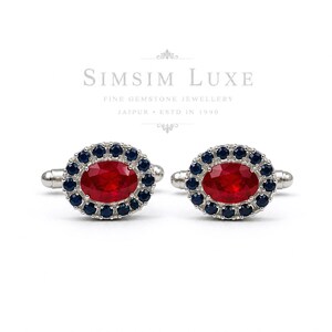 Puede incluir: Un par de gemelos de plata con una piedra preciosa roja de corte ovalado en el centro, rodeada por un halo de pequeños zafiros redondos de color azul oscuro. El texto SIMSIM LUXE FINE GEMSTONE JEWELLERY JAIPUR ESTD IN 1990 aparece en gris arriba.