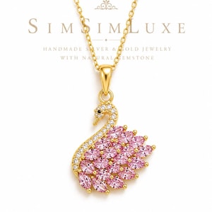 Può includere: Una collana color oro con un ciondolo a forma di cigno. Il collo del cigno è incastonato con piccole pietre chiare e il corpo è composto da pietre rosa taglio marquise. Il testo SIMSIMLUXE, HANDMADE SILVER & GOLD JEWELRY WITH NATURAL GEMSTONE e 925 STERLING SILVER è visibile.