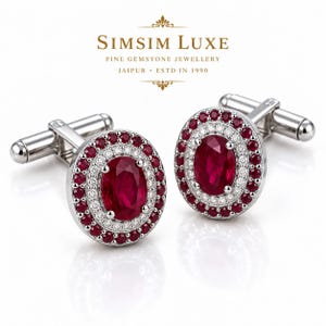 Puede incluir: Un par de gemelos plateados con una piedra central de rubí rojo intenso de corte ovalado, rodeada por un halo de pequeños diamantes blancos y un borde exterior de rubíes redondos. El texto SIMSIM LUXE FINE GEMSTONE JEWELLERY JAIPUR ESTD IN 1990 es visible.