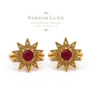 Może przedstawiać: Para złotych spinek do mankietów z motywem gwiazdy i okrągłym czerwonym kamieniem szlachetnym pośrodku. Tekst SIMSIM LUXE FINE GEMSTONE JEWELLERY JAIPUR ESTD IN 1990 jest wydrukowany powyżej ciemną czcionką na białym tle.
