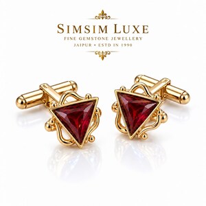 Op de afbeelding: Een paar gouden manchetknopen met driehoekige, gefacetteerde donkerrode edelstenen in sierlijke, gepolijste gouden monturen. Boven de manchetknopen staat de tekst SIMSIM LUXE FINE GEMSTONE JEWELLERY JAIPUR ESTD IN 1990 met decoratieve krullen.