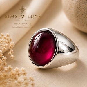 May include: A sterling silver 92.5 ring featuring a large, smooth, oval-shaped deep red gemstone set in a polished metal band. The text SIMSIM LUXE is visible in the upper left corner, along with the words Sterling Silver 92.5 and Timeless Elegance.