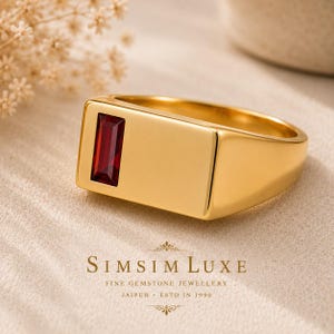 May include: A gold signet ring featuring a rectangular, deep red baguette-cut gemstone set on the left side of the flat, polished surface. The ring has a smooth, solid gold band and a minimalist design, with the text SIMSIM LUXE FINE GEMSTONE JEWELLERY JAIPUR ESTD IN 1990 printed below.