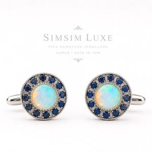 Op de afbeelding: Een paar ronde zilveren manchetknopen met een centrale iriserende opaal cabochon omringd door een halo van kleine, diepblauwe saffieren. De tekst SIMSIM LUXE FINE GEMSTONE JEWELLERY JAIPUR ESTD IN 1996 staat boven de manchetknopen.