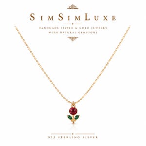 May include: A gold-toned chain necklace featuring a flower-shaped pendant with a round red gemstone center and two marquise-cut green gemstone leaves. The text SimSimLuxe, Handmade Silver & Gold Jewelry With Natural Gemstone, and 925 Sterling Silver appear.