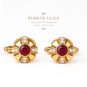 Puede incluir: Un par de gemelos dorados con un rubí rojo redondo central rodeado de pequeñas piedras blancas transparentes en un patrón radial. El texto SIMSIM LUXE FINE GEMSTONE JEWELLERY JAIPUR ESTD IN 1990 aparece impreso encima en letras oscuras.