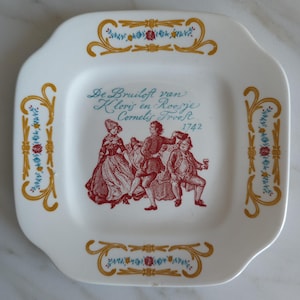 Puede incluir: Un plato cuadrado de cerámica blanca con una ilustración roja de tres figuras con vestimenta del siglo XVIII. El texto "De Bruiloft van Kloris en Roosje Cornelis Troost 1742" aparece en azul. El borde está decorado con volutas doradas y motivos florales.