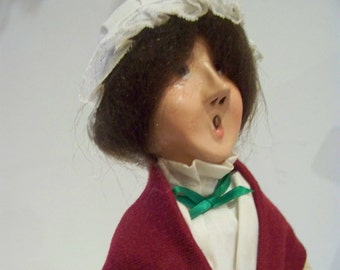May include: A vintage doll with a white lace bonnet, brown hair, and a red shawl. The doll has a painted face with a surprised expression and is wearing a white blouse with a green bow.