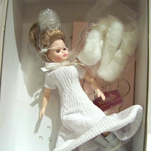 May include: A doll in a white dress with a full skirt, packaged in a box. The doll has blonde hair and is wearing white boots with bows. The box contains additional white accessories and a tag that reads "Narnia".