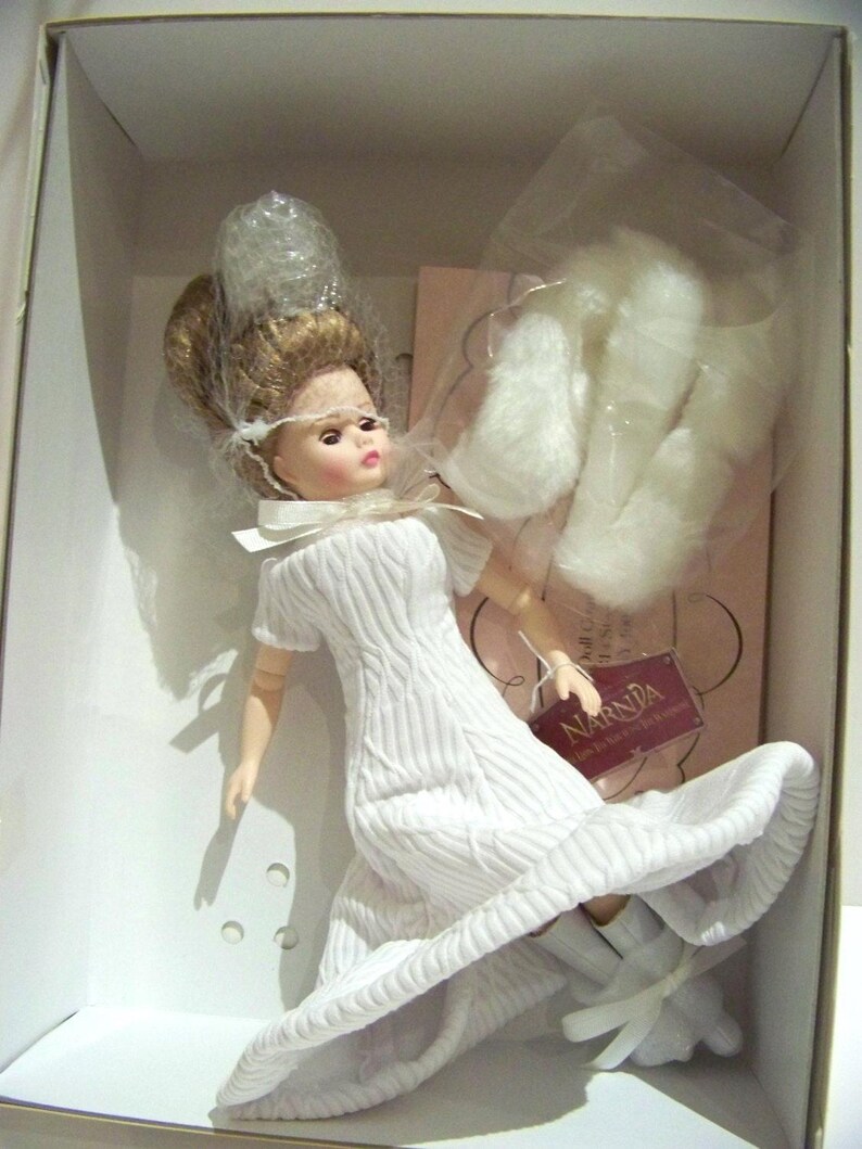 May include: A doll in a white dress with a full skirt, packaged in a box. The doll has blonde hair and is wearing white boots with bows. The box contains additional white accessories and a tag that reads "Narnia".