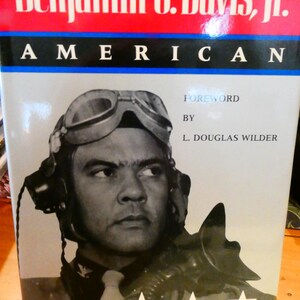 Può includere: Copertina di un libro con un ritratto in bianco e nero di Benjamin O. Davis, Jr., con casco e occhiali da aviatore. Il titolo "Benjamin O. Davis, Jr. AMERICAN" è in bianco su sfondo rosso e blu. Sono visibili tre stelle bianche.