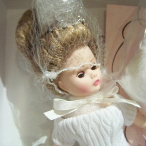 May include: A doll with blonde hair and a white dress with a textured knit pattern. The doll wears a white veil and a bow at the neckline. The doll is presented in a box.