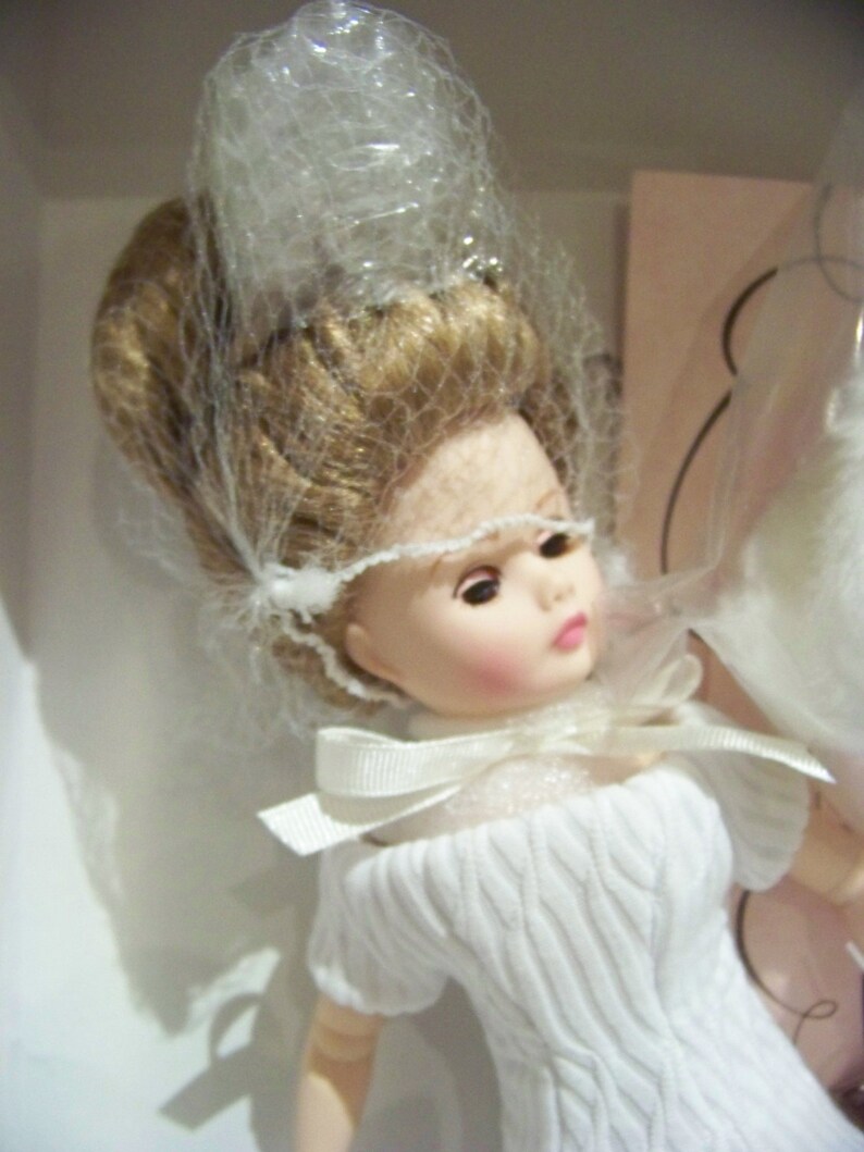 May include: A doll with blonde hair and a white dress with a textured knit pattern. The doll wears a white veil and a bow at the neckline. The doll is presented in a box.