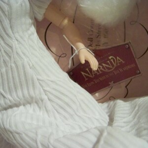 May include: A white doll in a textured dress, with a tag reading "NARNIA The Lion, The Witch, and The Wardrobe". The doll is in a box, with a white tag attached to its hand.
