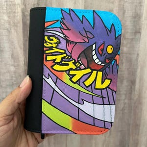 May include: A rectangular wallet featuring a black fabric spine and a vibrant graphic print on the front. The design displays a purple cartoon creature with a yellow eye and white teeth, accompanied by yellow Japanese text on a blue background.