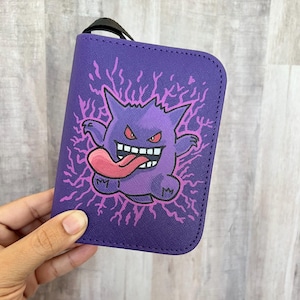 May include: A purple wallet featuring a graphic illustration of a round, spiky purple creature with red eyes, sharp white teeth, and a long pink tongue extended outward. The creature is surrounded by jagged pink lightning bolt patterns.