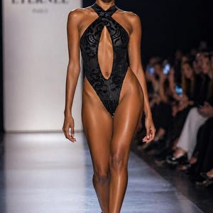 May include: A black bodysuit with a halter neckline and a keyhole cutout, featuring a subtle floral pattern. The model is wearing black strappy heels. The background includes the text "NOIR ÉTERNEL PARIS".