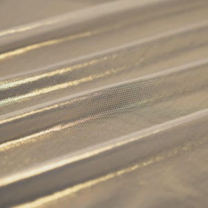 May include: A shimmering gold metallic fabric featuring a fine, semi-sheer mesh texture. The material is draped in soft, diagonal folds, creating a reflective surface that catches the light across its intricate, woven, and lightweight structure.