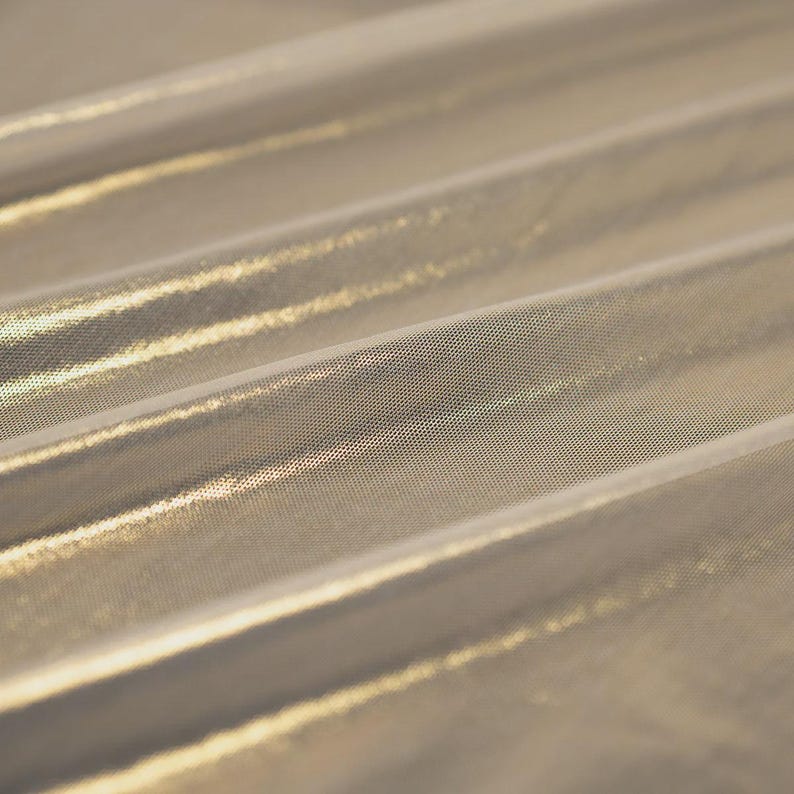 May include: A shimmering gold metallic fabric featuring a fine, semi-sheer mesh texture. The material is draped in soft, diagonal folds, creating a reflective surface that catches the light across its intricate, woven, and lightweight structure.