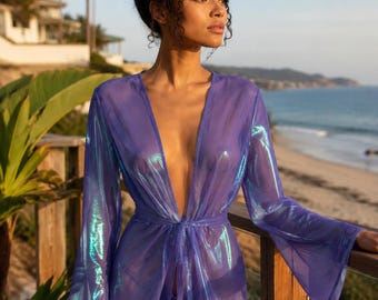 May include: A sheer, iridescent purple cover-up featuring long, flared sleeves, a deep V-neckline, and a matching tie-waist closure. The lightweight fabric displays a shimmering, holographic finish that reflects light with blue and green tones.