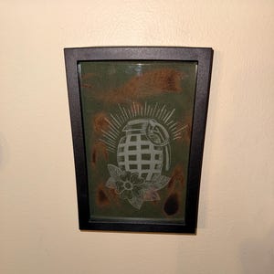 Puede incluir: Obra de arte enmarcada con un diseño de granada blanca, una flor y líneas radiantes sobre un fondo verde. El marco negro está montado en una pared beige. El diseño tiene una estética inspirada en los tatuajes.