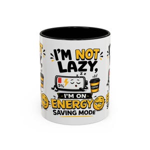 Peut inclure: Un mug en céramique blanc avec un intérieur et un bord noirs, orné d'un dessin animé représentant une batterie faible à 5 % avec les yeux fermés et des symboles Zz, à côté d'une tasse de café. Le texte indique I'M NOT LAZY, I'M ON ENERGY SAVING MODE.