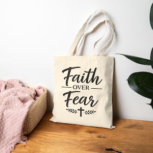 Peut inclure: Un sac fourre-tout en toile de couleur crème avec le texte imprimé en noir Faith OVER Fear, accompagné d'un petit symbole de croix et de motifs de feuilles décoratives. Le sac possède deux longues anses et une forme rectangulaire.