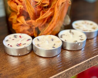 Peut inclure: Quatre petites bougies rondes dans des supports en métal argenté sont disposées sur une surface en bois. Les bougies blanches sont décorées de fleurs et d'herbes séchées. Un bocal en verre rempli de fleurs séchées orange est en arrière-plan.