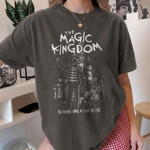 May include: A charcoal gray short-sleeve t-shirt featuring a white graphic print. The design includes the text THE MAGIC KINGDOM above a silhouette of a marching band member holding hands with a mouse character, with the text MY FATHER TOOK ME INTO THE CITY below.