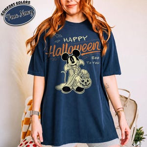 May include: A navy blue short-sleeve t-shirt featuring a graphic of a cartoon mouse dressed as a vampire holding a pumpkin bucket. The text reads HAPPY Halloween BOO TO YOU. A small oval logo in the top left corner says COMFORT COLORS True Navy.