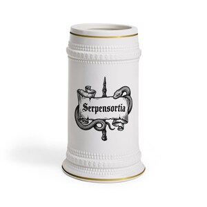 May include: A white ceramic beer stein featuring a gold rim at the top and bottom, with a textured beaded band near the base and rim. The centre displays a black ink illustration of a snake coiled around a wand and a scroll labelled Serpensortia.