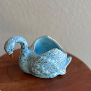 May include: A light blue ceramic swan-shaped planter or trinket dish featuring a delicate gold floral pattern across its body. The swan has a curved neck, a brown beak, and a gold-trimmed rim around the open top section of its back.