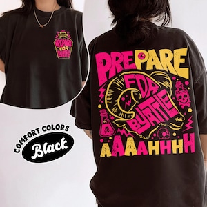 May include: A black t-shirt featuring a graphic design in pink and yellow. The front shows a small potion bottle with the text "PREPARE FOR BATTLE". The back displays a large ram head illustration with the text "PREPARE FOR BATTLE AAAAAHHHH".