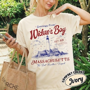 May include: An ivory t-shirt featuring a graphic print with the text Greetings From Widow's Bay, EST. 1681, MASSACHUSETTS, and The Next Martha's Vineyard. The illustration depicts a lighthouse, coastal buildings, clouds, and small stars.