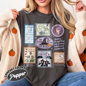 May include: A charcoal gray t-shirt featuring a collage of vintage-style postage stamp graphics with Halloween themes. The designs include a witch's house, a witch hat, and text such as "Always spilled salt" and "There's a little witch in all of us."