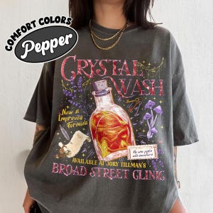 May include: A charcoal gray t-shirt featuring a graphic print of a glass bottle filled with orange liquid, surrounded by lavender, mushrooms, and a scroll. Text includes "Crystal Wash," "New & Improved Formula," and "He who fights with monsters."