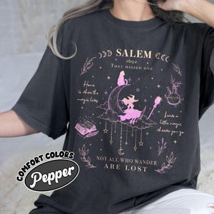 May include: A charcoal gray t-shirt featuring a pink and white graphic design with a crescent moon, a witch on a broomstick, a cat, a potion bottle, and an open book. Text includes SALEM 1692, THEY MISSED ONE, and NOT ALL WHO WANDER ARE LOST.