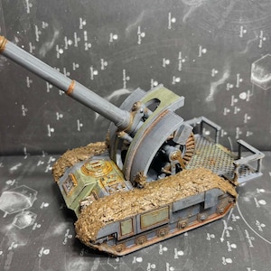 May include: A gray scale model of a self-propelled artillery vehicle featuring a long barrel, a large circular gear mechanism, and a flat rear platform. The tracks are covered in a textured brown mud-like material with metallic accents.