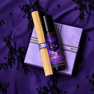 May include: A dark purple essential oil bottle with a black cap, a wooden stick, and a small gift box with a ribbon. The bottle has a cat graphic and the text "Gothic Moon Ritual Oils Velvet Crown Orange Vanilla Glamour". The background is a purple lace fabric.