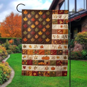 May include: A rectangular garden flag featuring a quilt-style design that mimics the American flag. The canton is dark brown with embroidered maple leaves in yellow, orange, and green. The stripes alternate between cream fabric and patchwork squares with autumn motifs, including pumpkins and foliage.