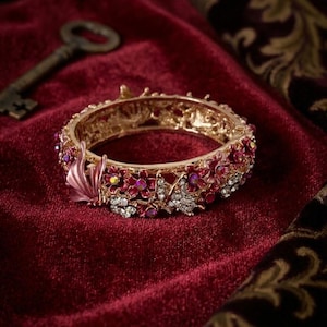 May include: A gold-tone hinged bangle bracelet featuring an intricate floral and butterfly motif. The design is embellished with pink enamel flowers, clear sparkling crystals, and a prominent pink butterfly accent on the side of the metal band.